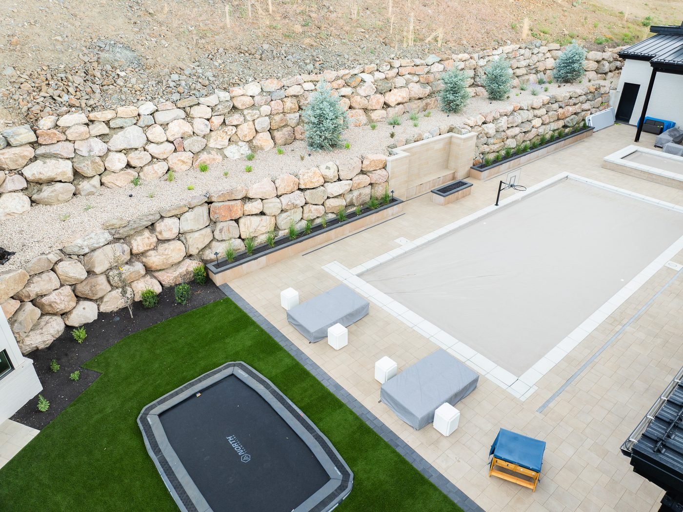 Aerial view of a paver pool deck with a contrasting border band, artificial turf lawn and tiered boulder retaining walls