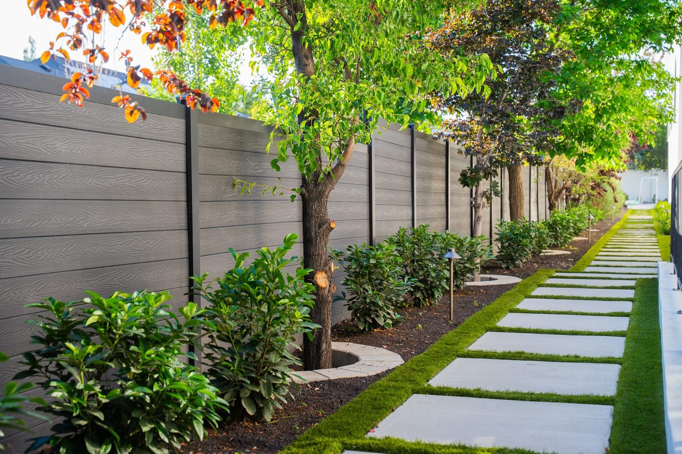 Paver stepping-stone path through planting beds alongside a fence line and young trees