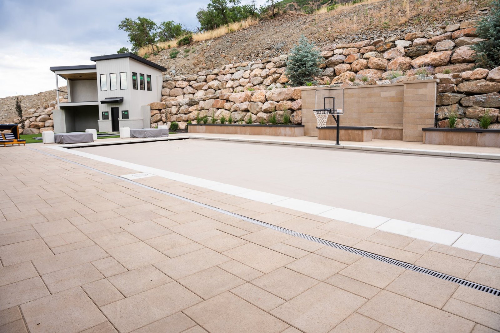 Large paver deck laid in running bond with a contrasting soldier-course border and a linear trench drain, beside a pool and tiered boulder retaining walls