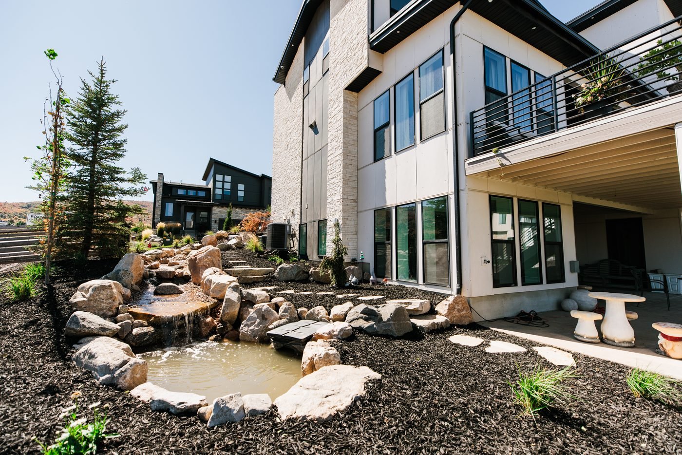Boulder waterfall spilling into a pond with stone stepping slabs beside a modern home