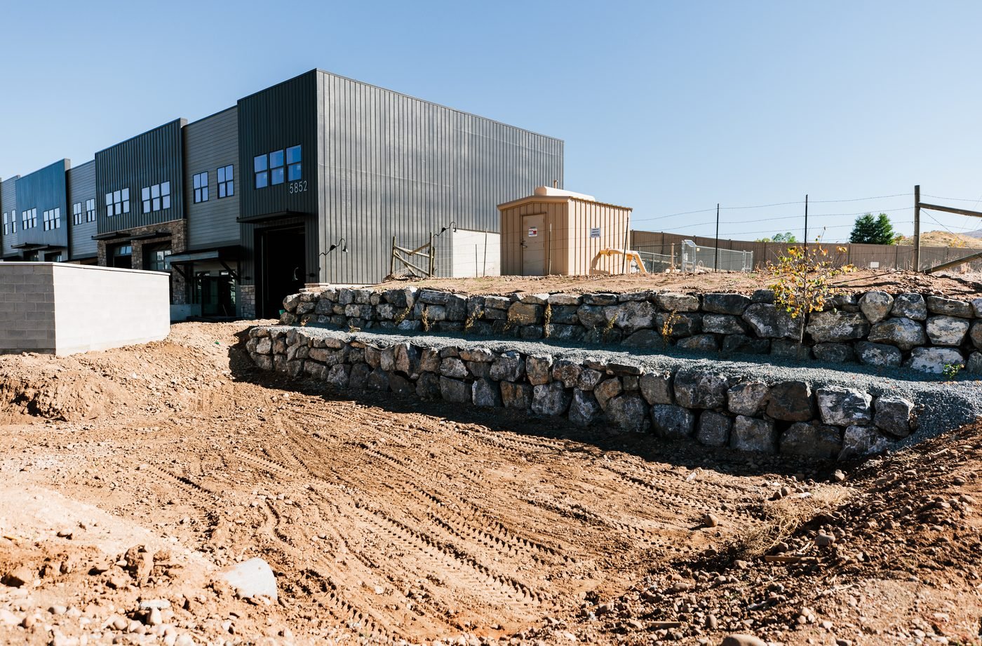 Graded commercial subgrade with machine tracks, beside two tiers of boulder retaining wall and drainage rock
